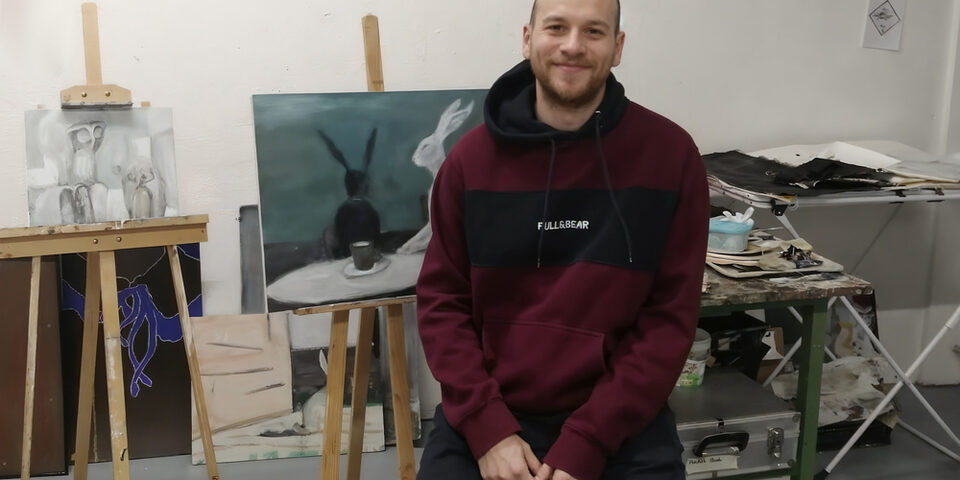 Eine Person in einem dunkelroten Pullover und dunkler Hose sitzt vor einer Staffelei auf welcher sich ein Bild befindet. Das Bild zeigt einen weißen und einen schwarzen Hasen an einem Tisch auf dem eine Tasse steht.