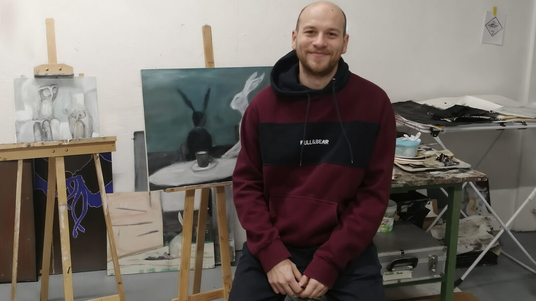 Eine Person in einem dunkelroten Pullover und dunkler Hose sitzt vor einer Staffelei auf welcher sich ein Bild befindet. Das Bild zeigt einen weißen und einen schwarzen Hasen an einem Tisch auf dem eine Tasse steht.