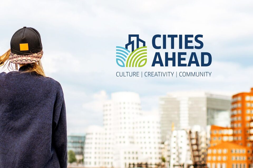 Junge Frau mit Cap von hinten, die auf eine Ansammlung von mehrstöckigen Häusern blickt. Darüber das "Cities Ahrad"-Logo