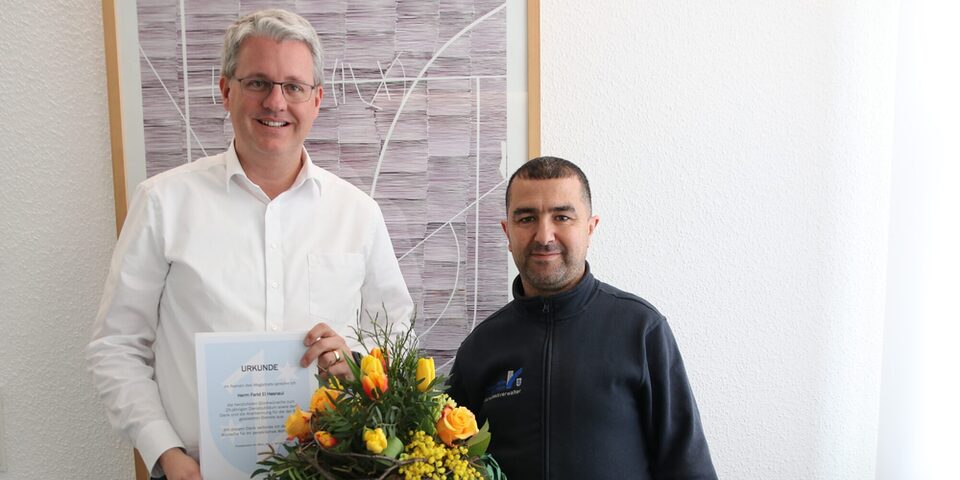 Der Oberbürgermeister zeigt die Urkunde und El Hasnaui hält einen Blumenstrauß in der Hand.