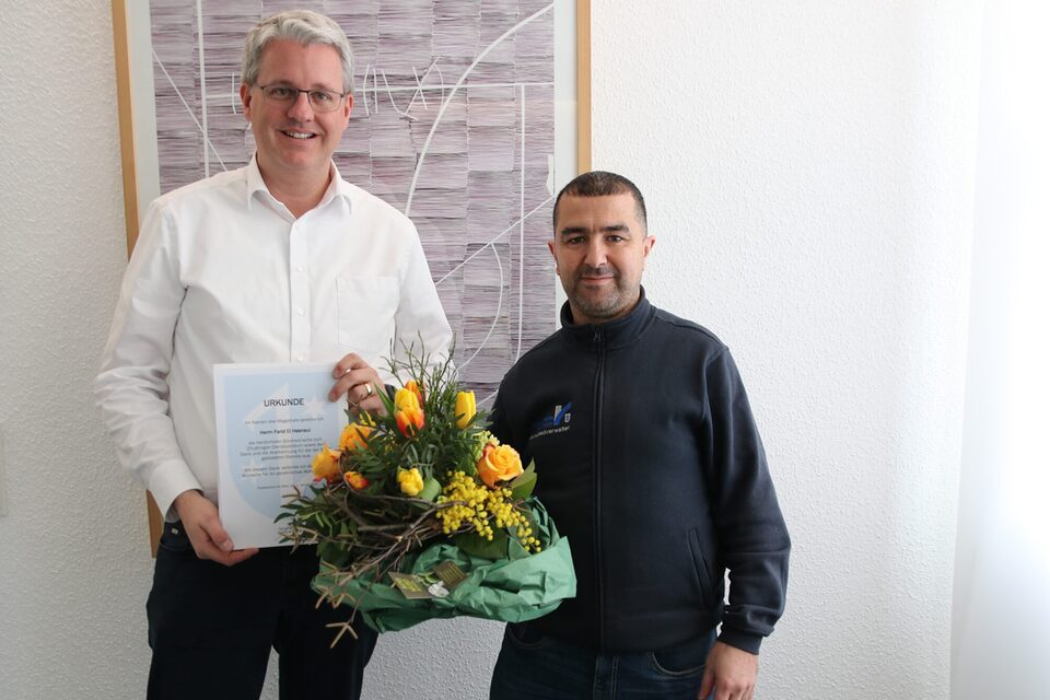 Der Oberbürgermeister zeigt die Urkunde und El Hasnaui hält einen Blumenstrauß in der Hand.