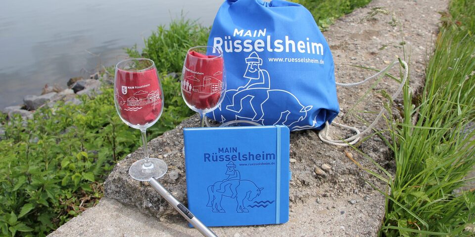 Zwei Weingläser, ein Notizbuch und ein Turnbeutel sind vor dem Main drapiert. Alle Produkte haben die Aufschrift "MAIN Rüsselsheim".