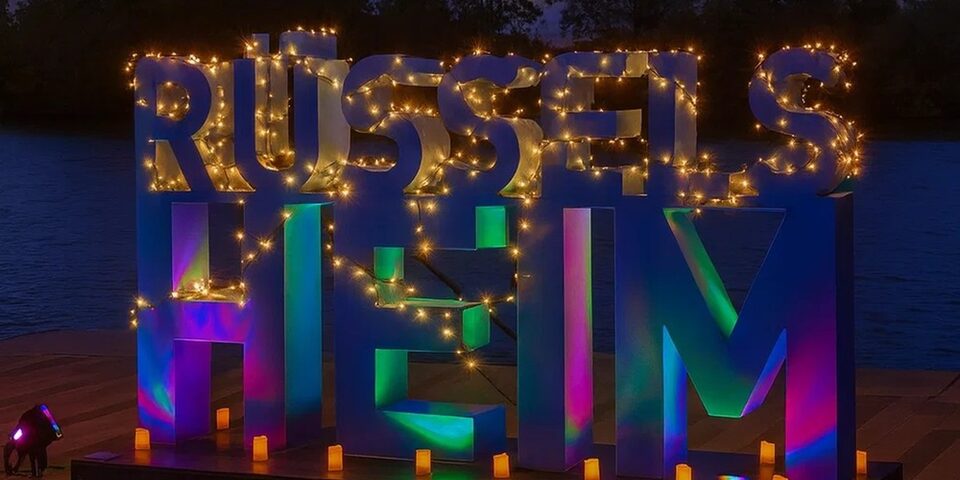 Rüsselsheim-Schriftzug, der mit Lichterketten beleuchtet ist und bunt schimmert. Im Hintergrund im Abenddunkel der Main.