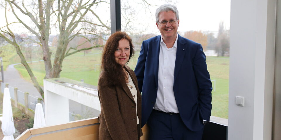 Dr. Beate Kemfert (Vorstand der Stiftung Opelvillen) und Oberbürgermeister Patrick Burghardt stehen vor einem Fenster in den Opelvillen mit Blick auf das grüne Mainvorland.
