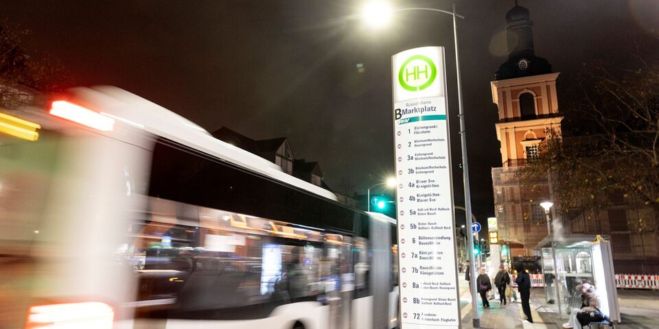 Bushaltestelle "Marktplatz" im Dunkeln. Ein vorbeifahrender Bus, wartende Menschen. Im Hintergrund die Stadtkirche.