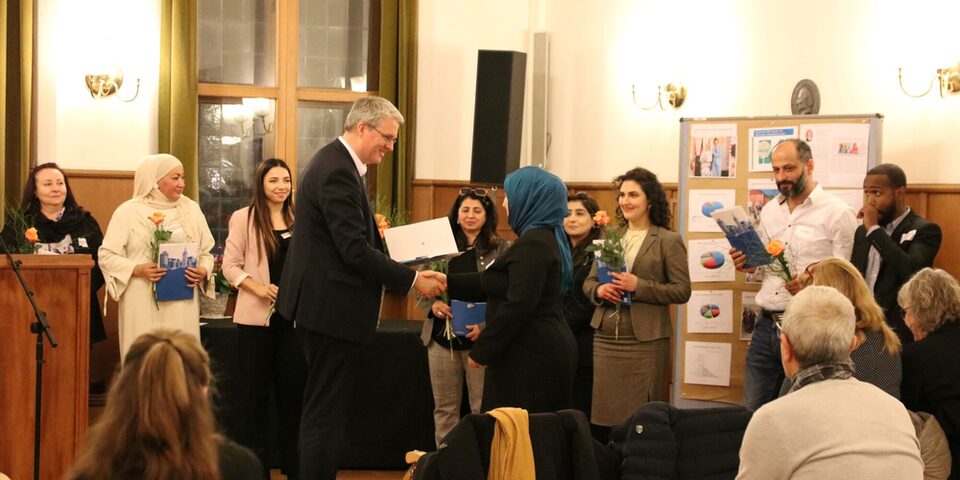 Oberbürgermeister Patrick Burghardt übergibt einer Dame die Urkunde für die Integrationslotsen-Qualifizierung im Trauzimmer. Im Hintergrund stehen bereits andere Integrationslotsinnen und -lotsin mit ihren Urkunden und einer orangenen Rose in der Hand.