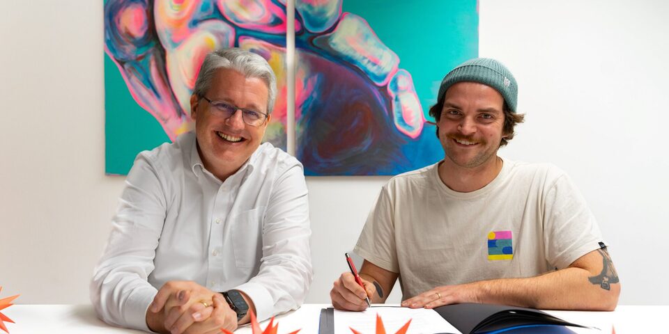 Oberbürgermeister Patrick Burghardt und Benny Becker (Rollwerk) sitzen an einem Tisch und lächeln in die Kamera. Benny Becker hält einen Stift, um die Fördervereinbarung mit der Stadt zu unterzeichnen, die vor ihm auf dem Tisch liegt.