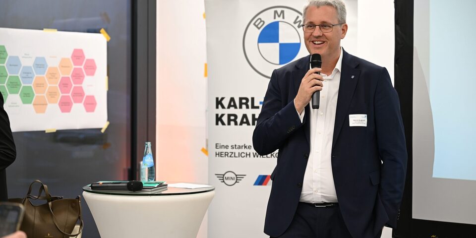 Oberbürgermeister Patrick Burghardt spricht lächelnd in ein Mikrofon. Im Hintergrund ein Rollup des Autohauses Karl+Co. sowie ein Stehtisch und mehrere Poster.