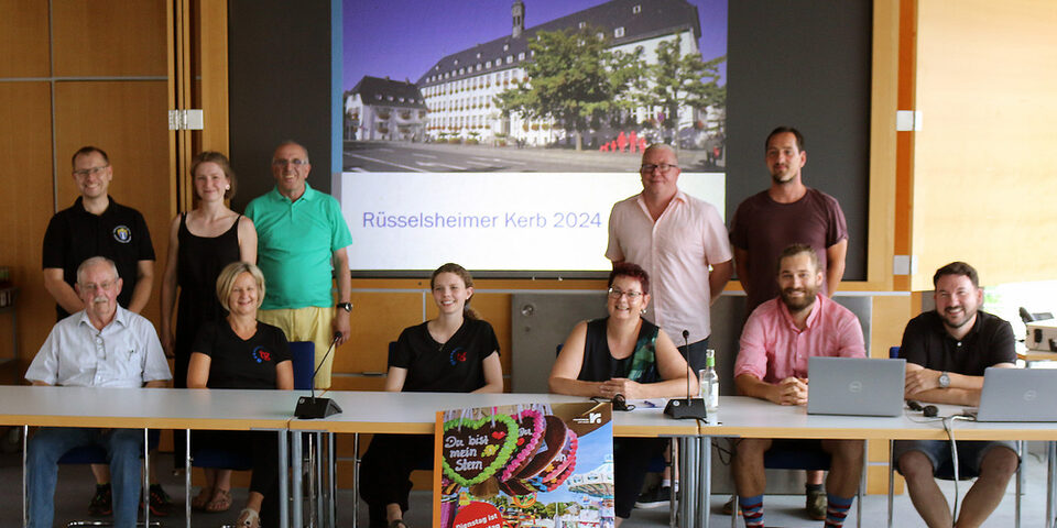 Das Team des Veranstaltungswesens der Stadt und Vereinsvertreterinnen und -vertreter haben sich vor einer Powerpoint-Präsentation mit der Aufschrift "Rüsselsheimer Kerb 2024" positioniert. Im Vordergrund das diesjährige Plakatmotiv.