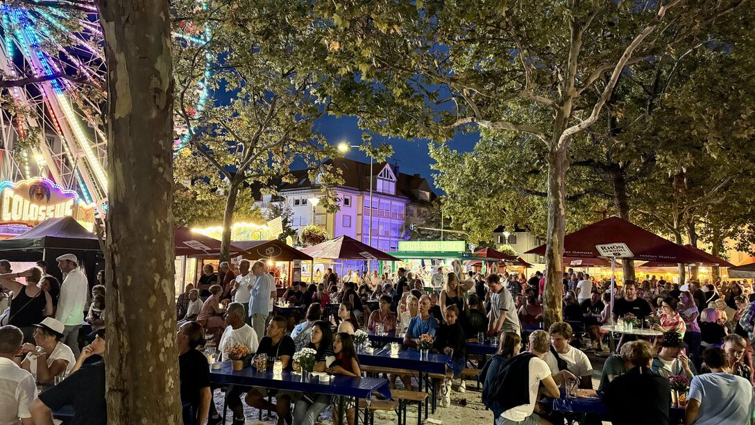 Gut gefüllte Sitzplätze im Kerbetreff bei Abendstimmung. Erleuchtetes Riesenrad und Buden im Hintergrund.