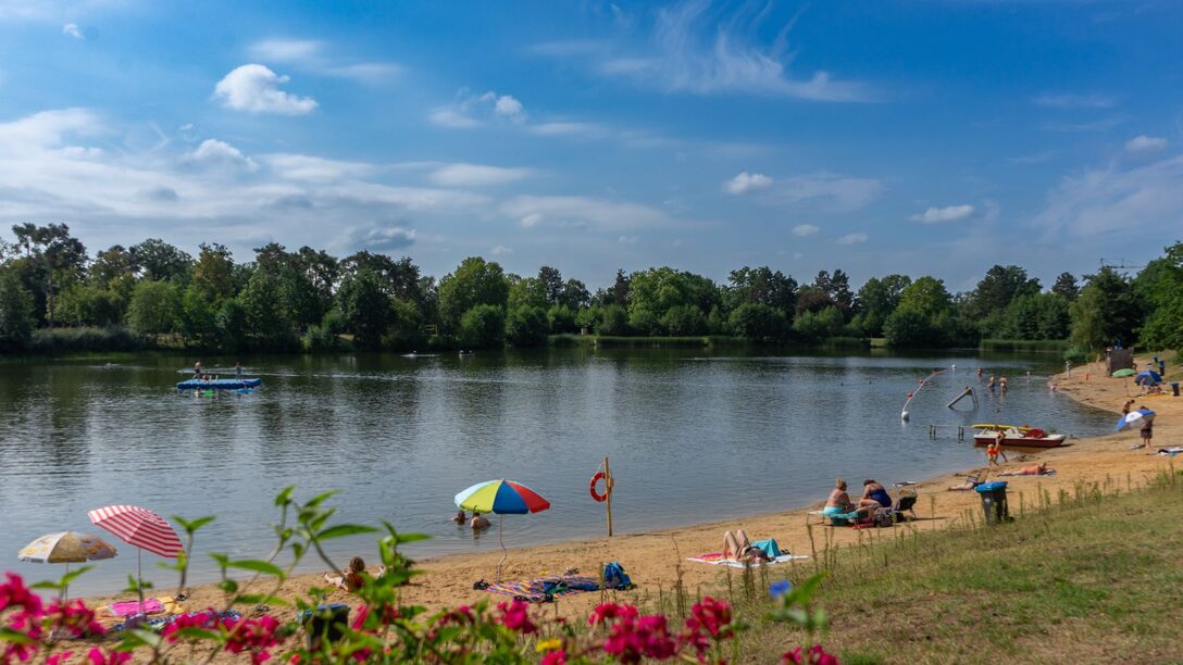 Sandstrand und Blick auf den See im Waldschwimmbad. Vereinzelt bunte Sonnenschirme und Liegeflächen.