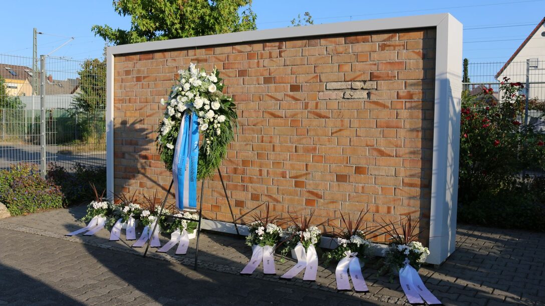 Mahnmal in der Grabenstraße: Mauer und ein großer Kranz mit Schleife sowie kleinere Kränze davor.