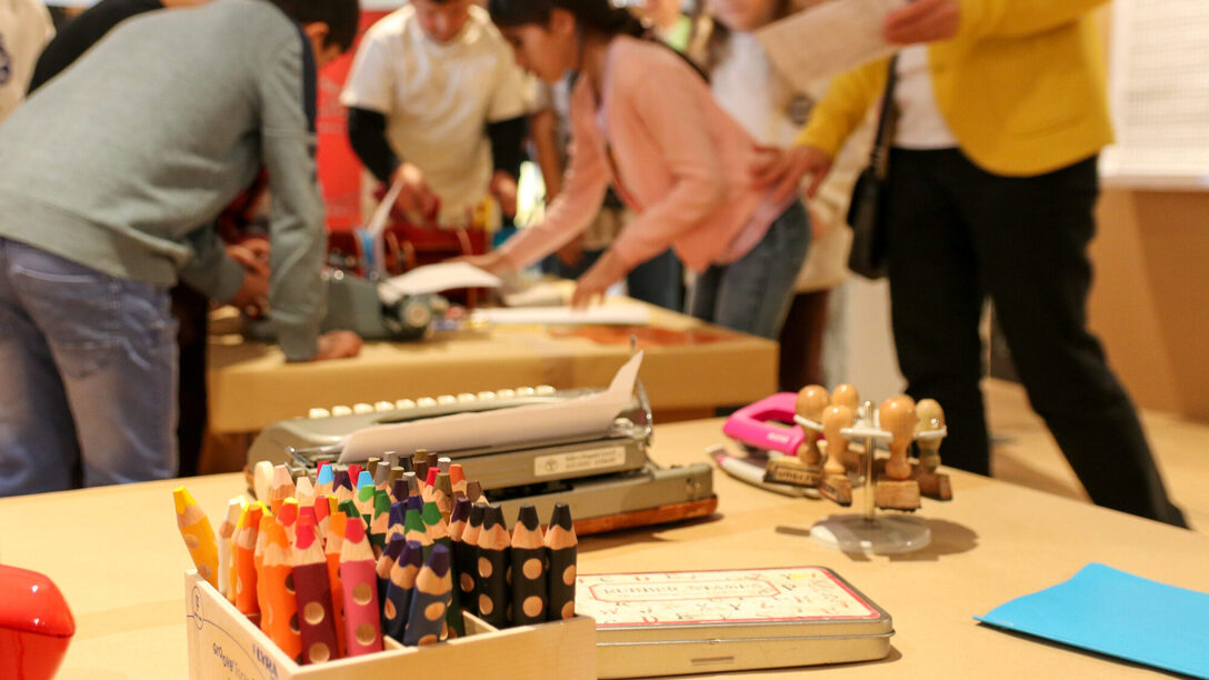 Ein Teil der Ausstellung Spielplatz Sprache. Im Vordergrund sind Buntstifte, eine Schreibmaschine, Stempel und ein Stempelkissen zu sehen. Im Hintergrund beugen sich mehrere Kinder über eine weitere Schreibmaschine.