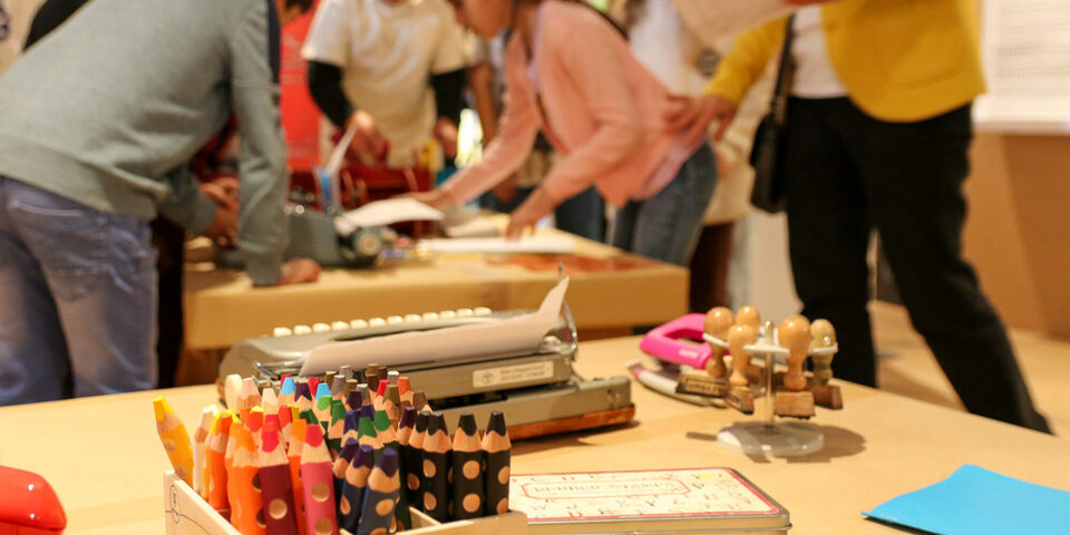 Ein Teil der Ausstellung Spielplatz Sprache. Im Vordergrund sind Buntstifte, eine Schreibmaschine, Stempel und ein Stempelkissen zu sehen. Im Hintergrund beugen sich mehrere Kinder über eine weitere Schreibmaschine.