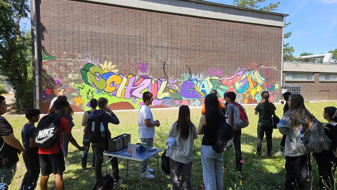 Eine Gruppe Jugendlicher steht vor einem Graffiti auf einer Gebäudewand. In bunten Buchstaben steht dort "Schulgarten"