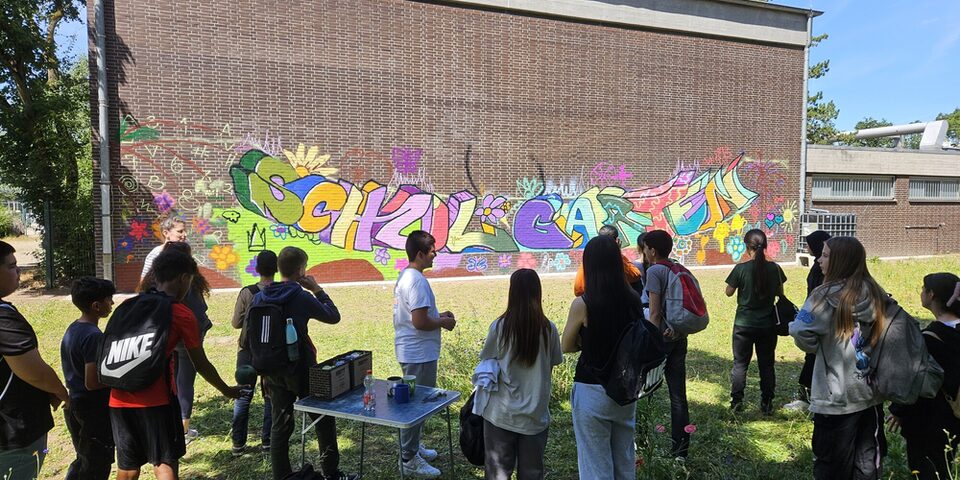 Eine Gruppe Jugendlicher steht vor einem Graffiti auf einer Gebäudewand. In bunten Buchstaben steht dort "Schulgarten"