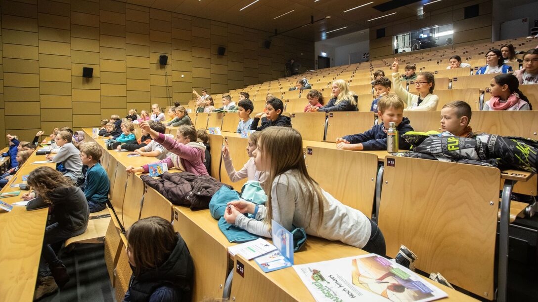 Kinder bei KinderUni in einem Hörsaal, die sich melden oder gespannt nach vorn blicken.