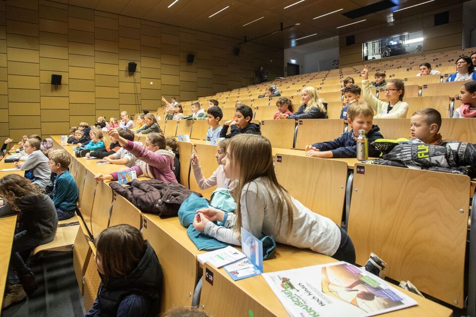 Kinder bei KinderUni in einem Hörsaal, die sich melden oder gespannt nach vorn blicken.