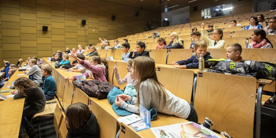 Kinder bei KinderUni in einem Hörsaal, die sich melden oder gespannt nach vorn blicken.