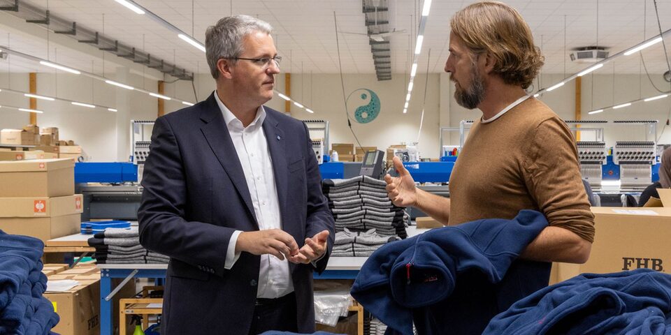 Oberbürgermeister Patrick Burghardt und Wailua-Geschäftsführer Mark Flynn gestikulierend im Gespräch vor bestickten Jacken. Kartons und Produktionshalle im Hintergrund.