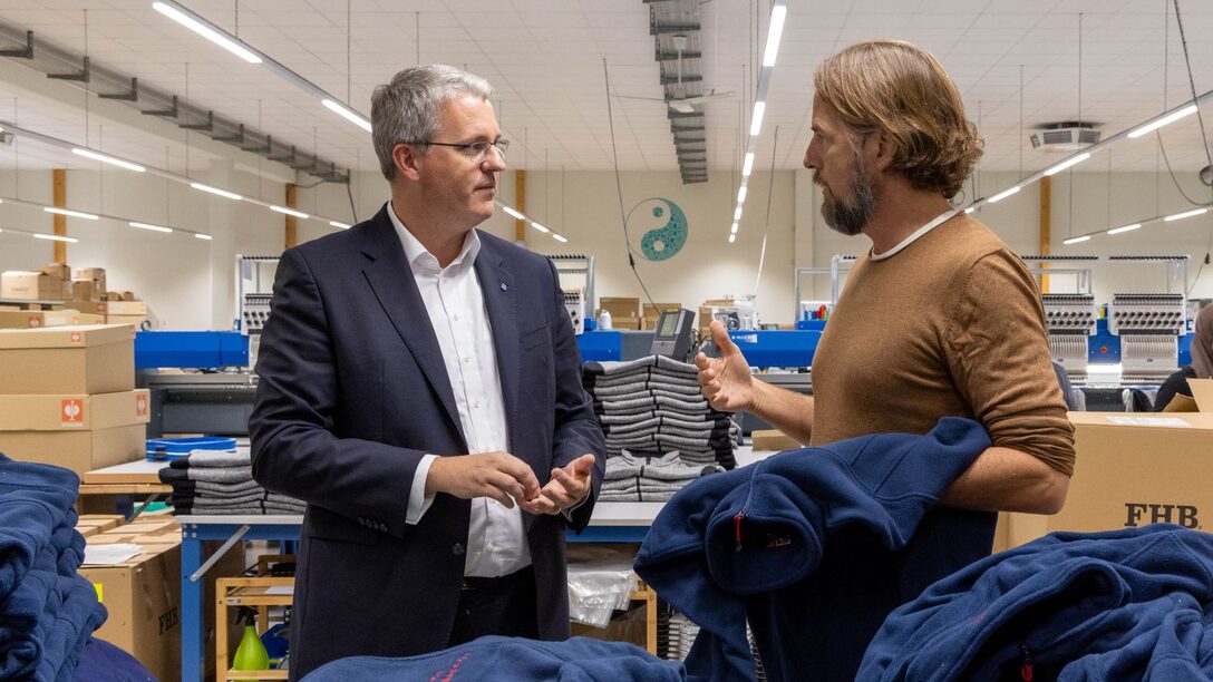 Oberbürgermeister Patrick Burghardt und Wailua-Geschäftsführer Mark Flynn gestikulierend im Gespräch vor bestickten Jacken. Kartons und Produktionshalle im Hintergrund.