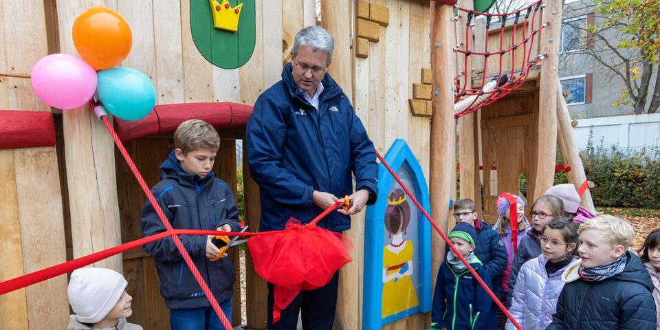 Oberbürgermeister Patrick Burghardt mit dem neunjährigen Emil, die gemeinsam ein rotes Band vor dem neuen Ritterburg-Klettergerät durchschneiden.