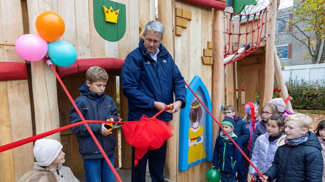 Oberbürgermeister Patrick Burghardt mit dem neunjährigen Emil, die gemeinsam ein rotes Band vor dem neuen Ritterburg-Klettergerät durchschneiden.