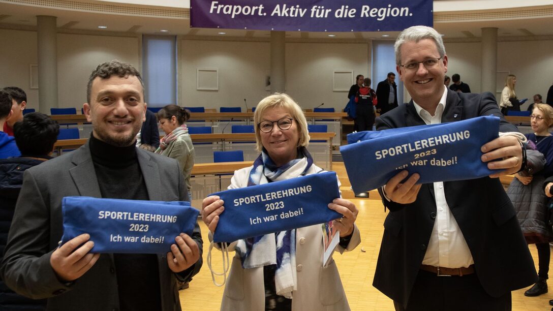 Die beiden stellvertretenden Stadtverordnetenvorsteher Yildiray Safel und Heide Böcker sowie Oberbürgermeister Patrick Burghardt halten blaue Turnbeutel mit der Aufschrift "Sportlerehrung 2023 - Ich war dabei" lächelnd in die Kamera.