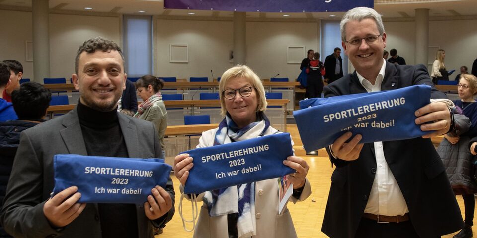 Die beiden stellvertretenden Stadtverordnetenvorsteher Yildiray Safel und Heide Böcker sowie Oberbürgermeister Patrick Burghardt halten blaue Turnbeutel mit der Aufschrift "Sportlerehrung 2023 - Ich war dabei" lächelnd in die Kamera.