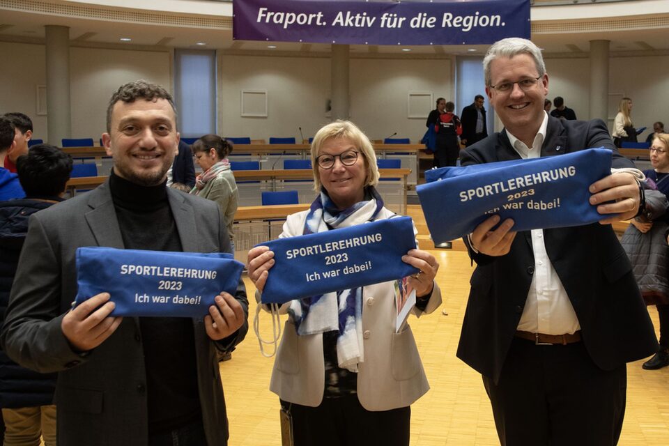 Die beiden stellvertretenden Stadtverordnetenvorsteher Yildiray Safel und Heide Böcker sowie Oberbürgermeister Patrick Burghardt halten blaue Turnbeutel mit der Aufschrift "Sportlerehrung 2023 - Ich war dabei" lächelnd in die Kamera.