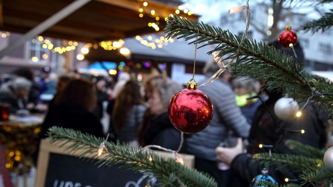 Rote Weihnachtskugel hängt an Tannenbaumzeig, der ins Bild ragt. Im Hintergrund sind verschwimmen Buden, Lichter und Menschen zu sehen.