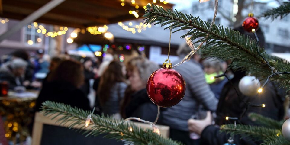 Rote Weihnachtskugel hängt an Tannenbaumzeig, der ins Bild ragt. Im Hintergrund sind verschwimmen Buden, Lichter und Menschen zu sehen.