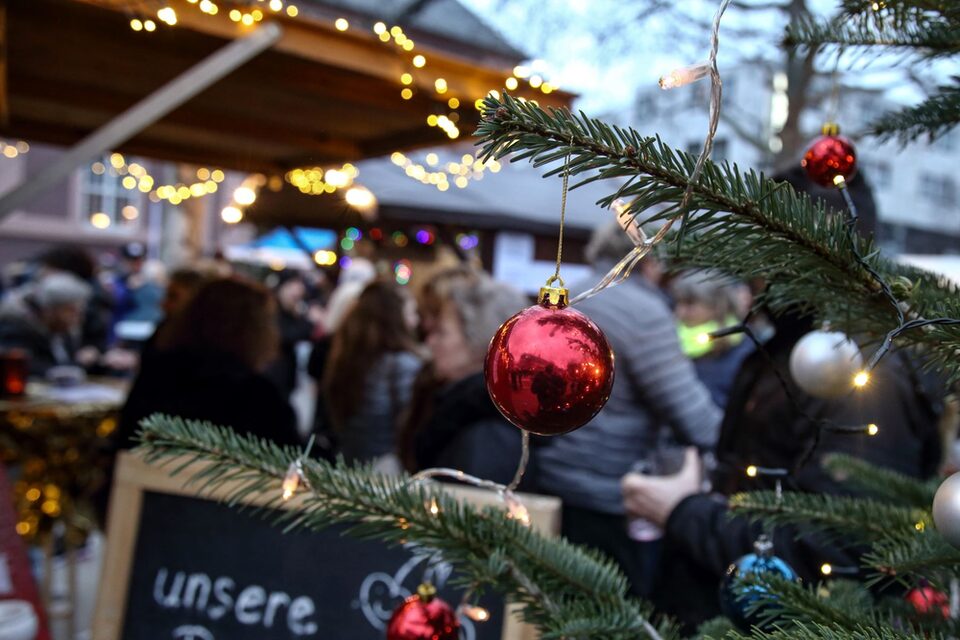 Rote Weihnachtskugel hängt an Tannenbaumzeig, der ins Bild ragt. Im Hintergrund sind verschwimmen Buden, Lichter und Menschen zu sehen.