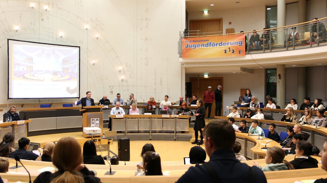 Bürgermeister Dennis Grieser steht am Rednerpult des mit Kindern und Jugendlichen gut gefüllten Ratssaals. An der Galerie hängt ein orangenes Banner mit der Aufschrift "Stadt Rüsselsheim. Jugendförderung".