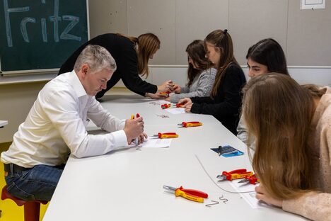 Schülerinnen und Mann in weißem Hemd sitzen an Tisch und probieren sich an Drähten und Zangen aus. Im Hintergrund eine Tafel mit der Aufschrift "Fritz".