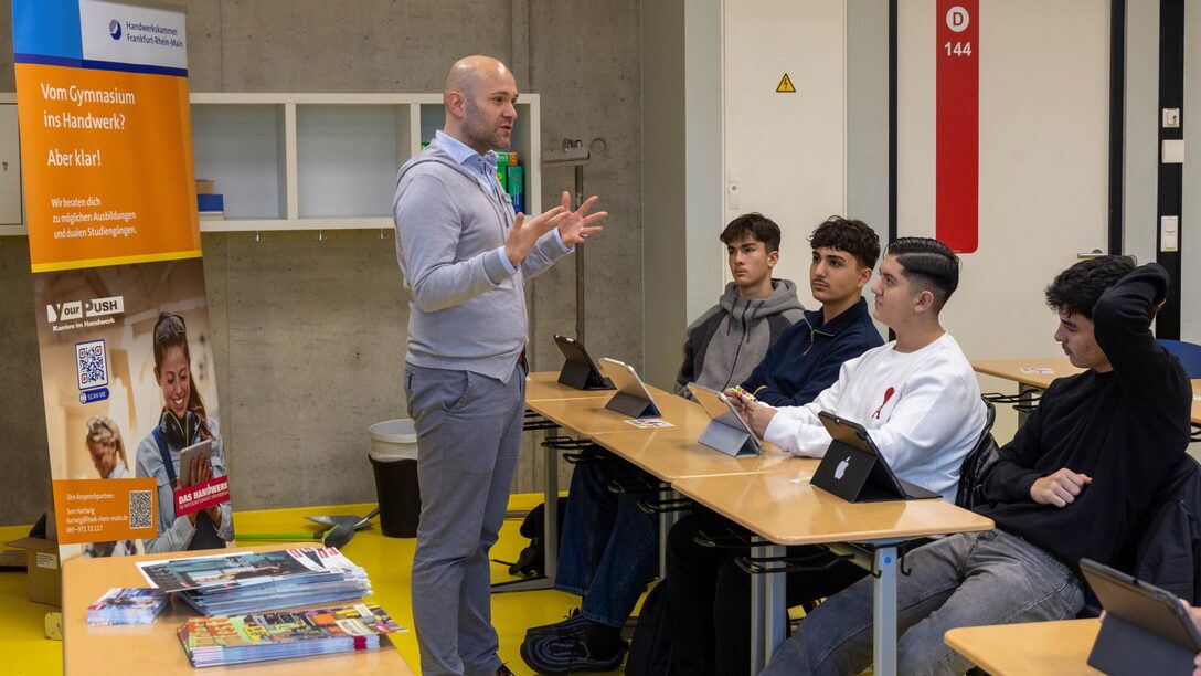 Links im Bild: Rollup der Handwerkskammer Frankfurt-Rhein-Main. In der Mitte ein Mann in grauer Weste und grauer Hose, der präsentiert und gestikuliert. Rechts eine Tischreihe, an denen Schüler sitzen und den Mann interessiert anschauen.