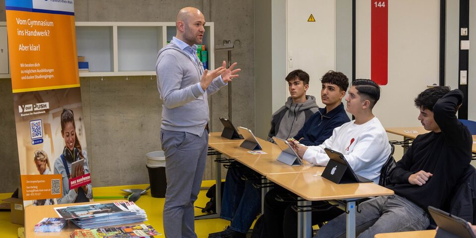 Links im Bild: Rollup der Handwerkskammer Frankfurt-Rhein-Main. In der Mitte ein Mann in grauer Weste und grauer Hose, der präsentiert und gestikuliert. Rechts eine Tischreihe, an denen Schüler sitzen und den Mann interessiert anschauen.