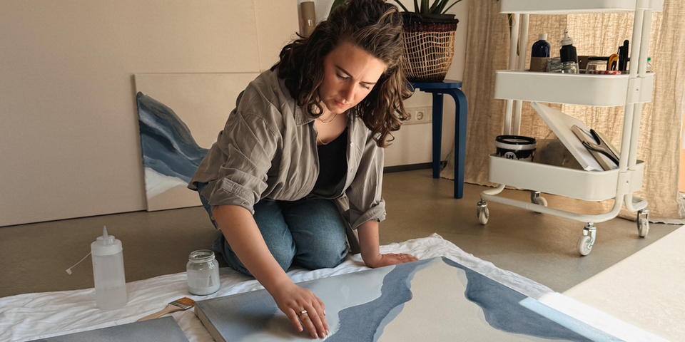 Eine Person kniet vor einer Leinwand auf der künstlerische Wellen in Blautönen und Weiß zu sehen sind