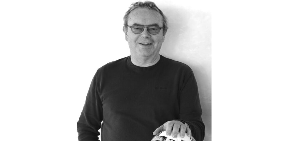 Schwarz-weißes Foto von Robert Neubauer, dessen linke Hand auf einem Fußball auf dem Tisch vor sich liegt.