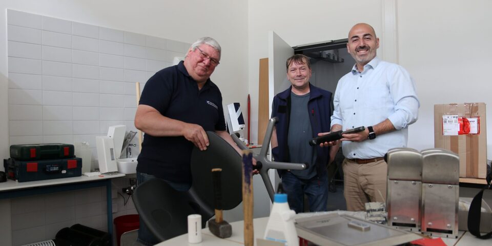 Hinter einem Tisch mit Werkzeug zeigen die Schulhausverwalter dem Bürgermeister einen Stuhl und die Druckluftfeder, die ausgetauscht werden musste.