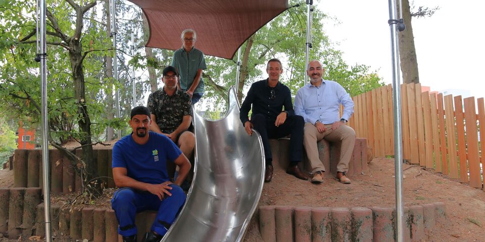 Stadtrat, Bürgermeister Mitarbeiter der Grünplanung und der Spielplatzcrew sitzen an der Rutsche unter dem Sonnersegel auf dem neu modulierten Spielhügel.