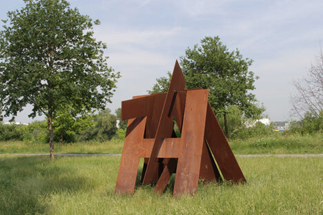 Foto: Stahl-Skulptur aus ineinander verschränkten Buchstaben steht auf einer Wiese