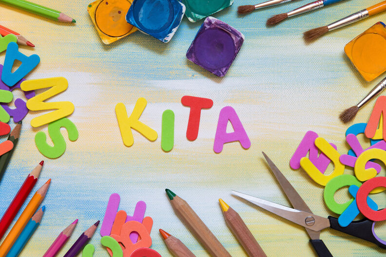 Wort Kita aus bunten Buchstaben gebastelt umgeben von Buntstiften, Schere und weiteren Buchstaben
