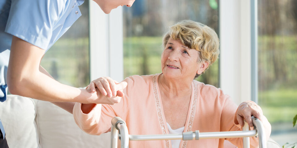 Senioren mit Gehhilfe wird unterstützt durch eine Pflegekraft