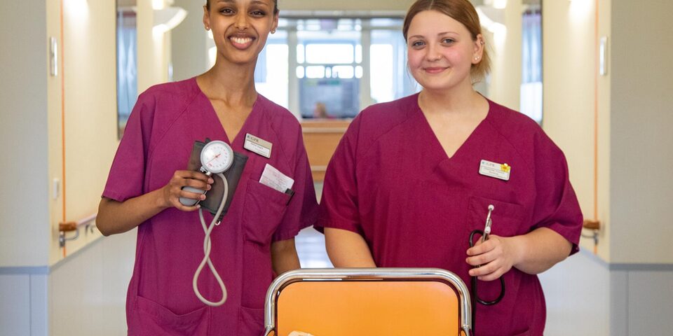 Zwei Pflegekräfte des GPR Klinikums stehen im Stationsgang und halten Blutdruckgerät und Stethoskop in der Hand