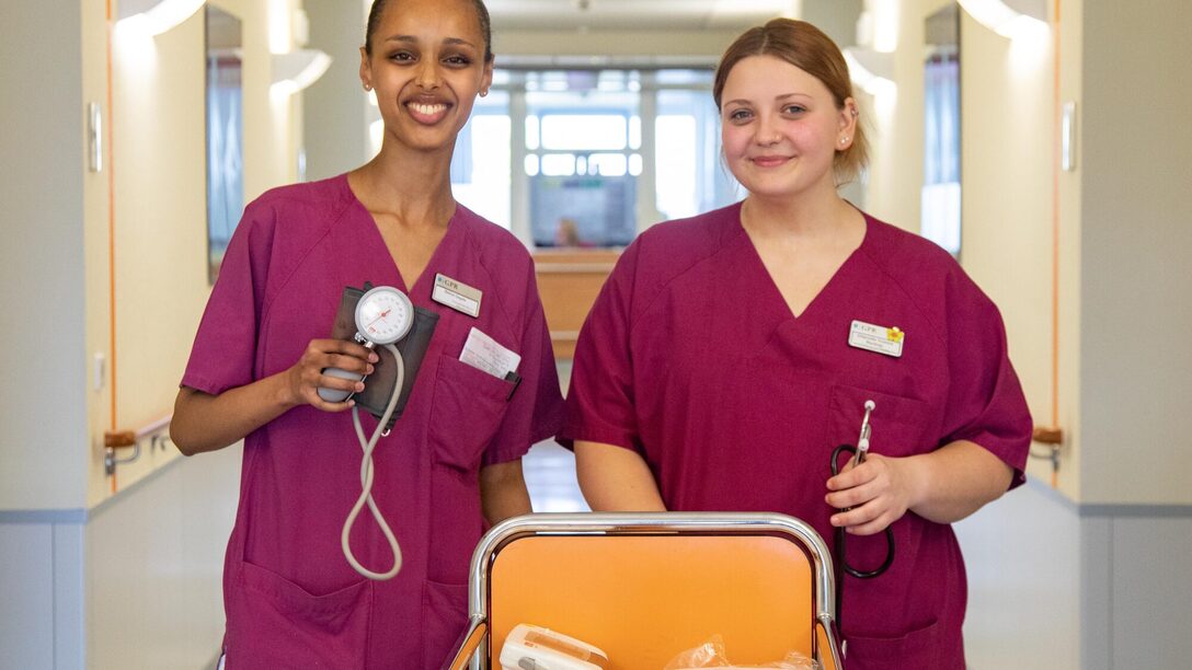 Zwei Pflegekräfte des GPR Klinikums stehen im Stationsgang und halten Blutdruckgerät und Stethoskop in der Hand