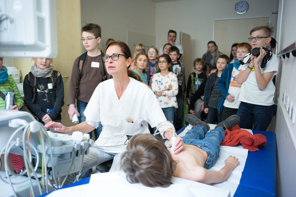 Kinder-Uni-Workshop: Ärztin im weißen Kittel macht eine Ultraschalluntersuchung zur Röntgenstrahlen-Vorlesung. Viele Kinder schauen gebannt zu.