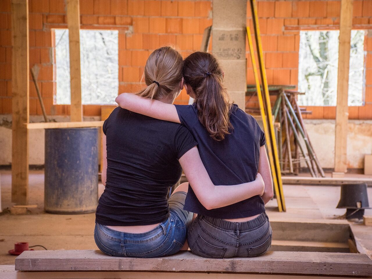 Zwei Frauen von hinten, die sich umarmen. Im Hintergrund ist eine Baustelle zu sehen.