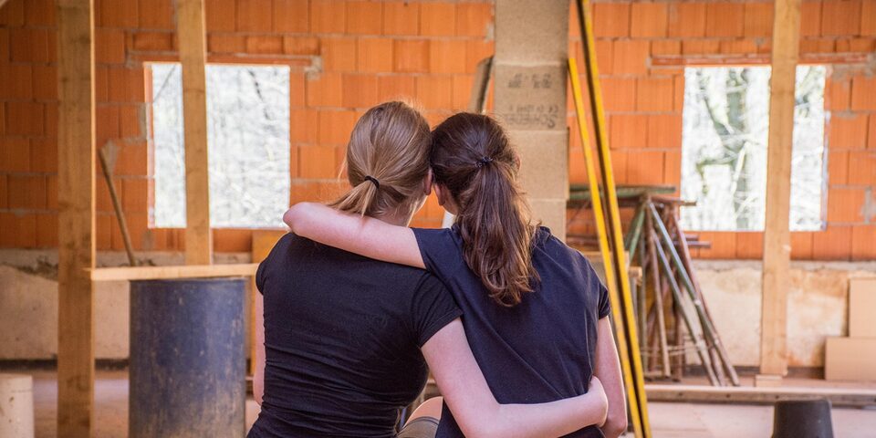 Zwei Frauen von hinten, die sich umarmen. Im Hintergrund ist eine Baustelle zu sehen.