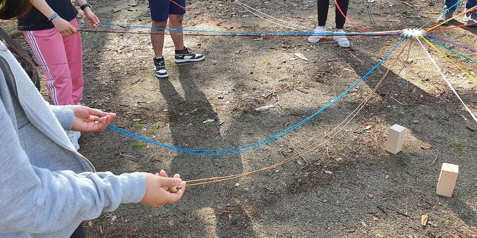 Jugendliche halten bunte Schnüre in den Händen, mit denen sie einen Holzklotz anheben.